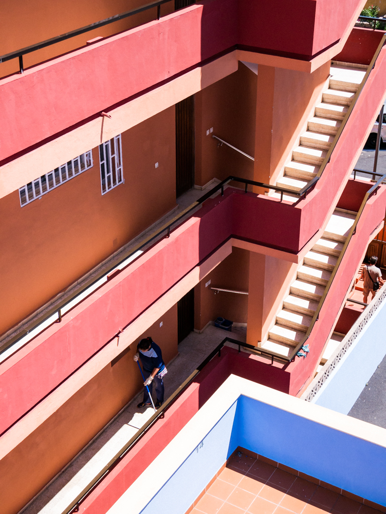 Femme passant le balai dans les escaliers d'un immeuble à Ténérife, se tenue bleu contraste avec les murs rouges et oranges