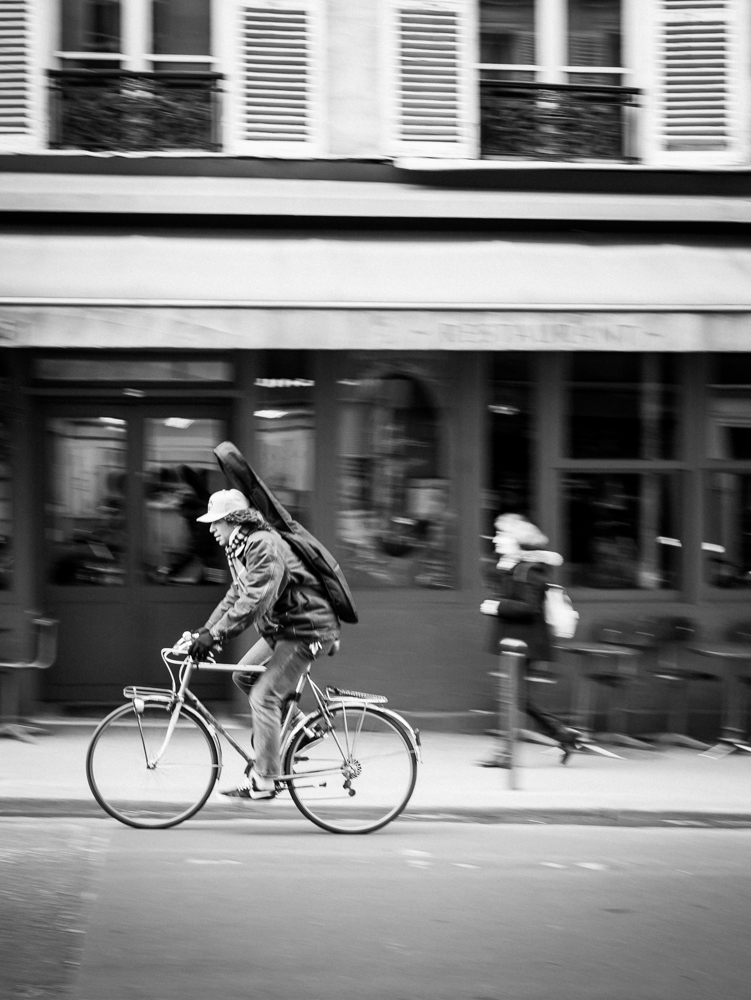Homme à vélo passe dans le trafic des rues de Paris, une guitare sur le dos